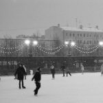 Black and white photo of people ice skating outdoors at night, under string lights in a winter setting.